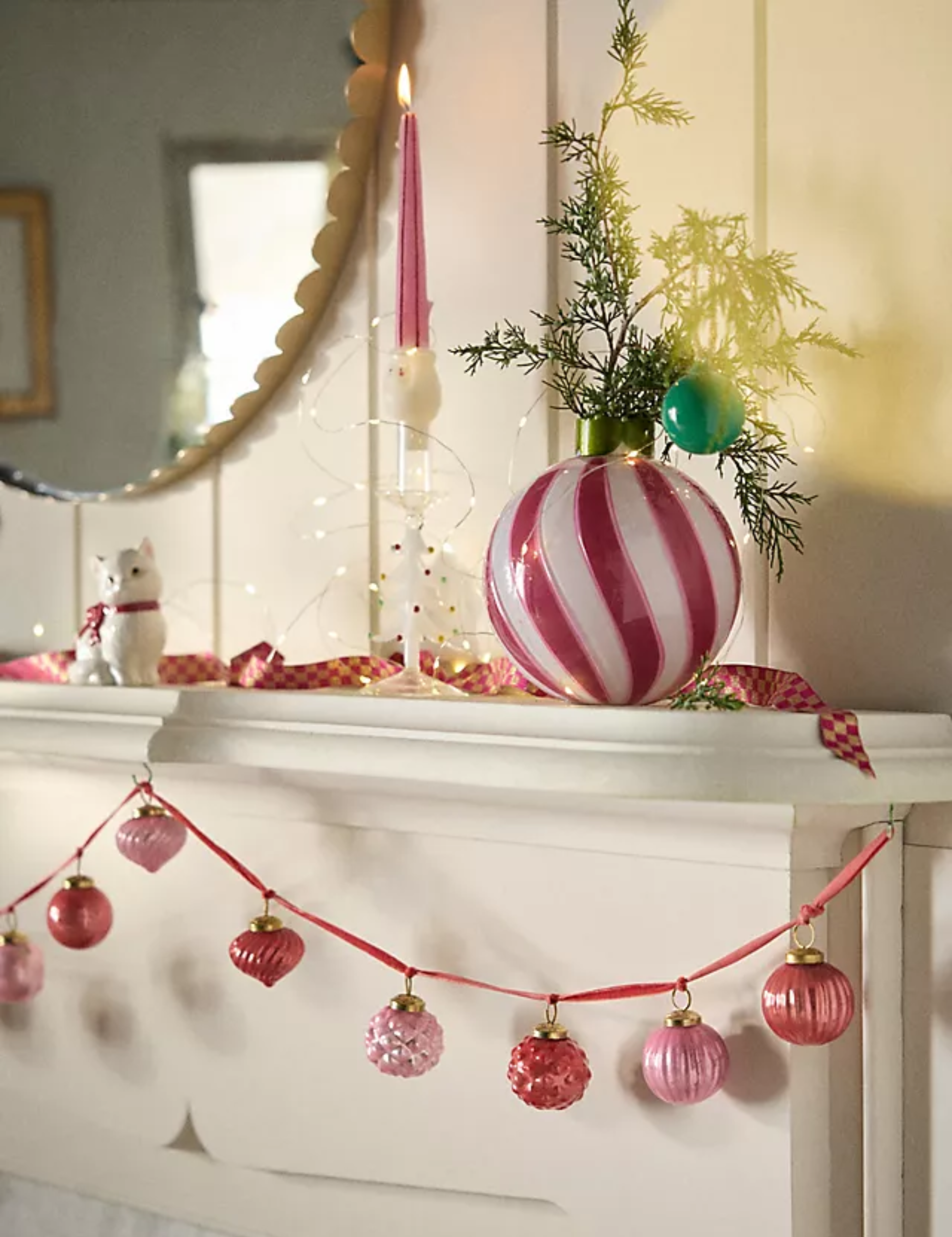 Ornament garland displayed on a white fireplace mantel topped with an oversized striped ornament, taper candle, and miniature fern tree