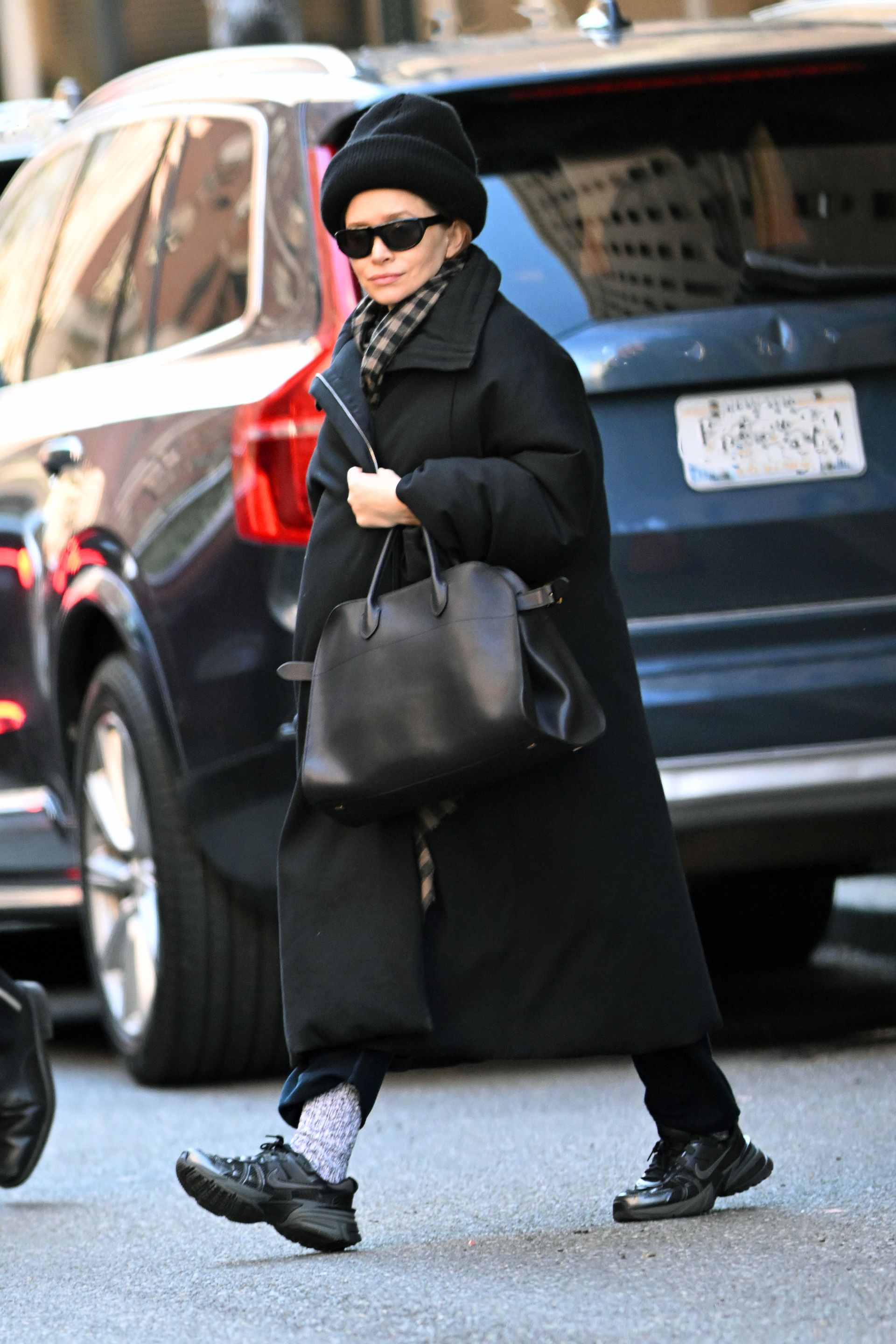 Ashley olsen wears a black hat, black sunglasses, plaid scarf, long black coat, gray socks, black the row margaux bag, and black v2k nike sneakers.