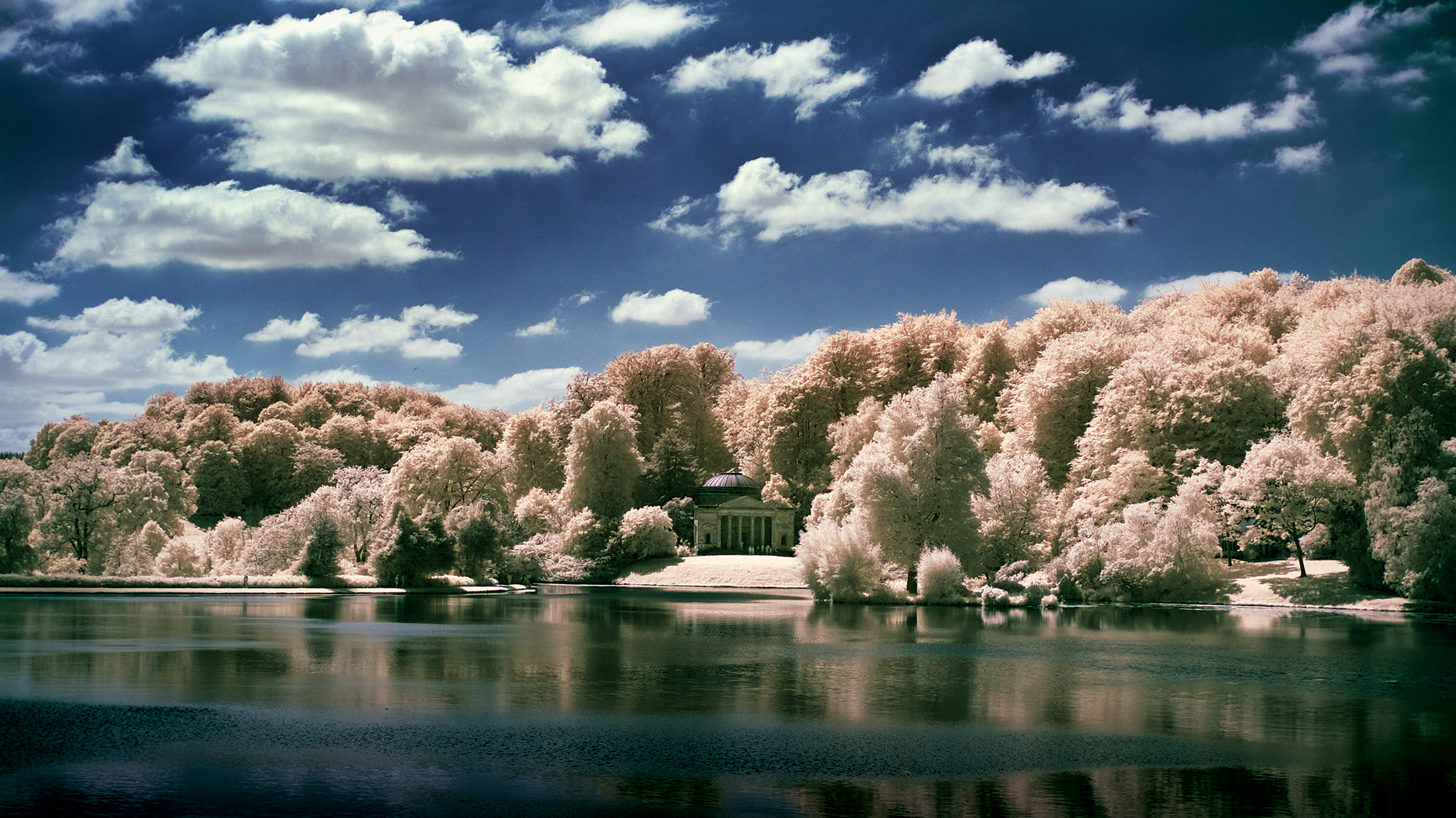 A serene lakeside with lush trees in soft pastel hues under a blue sky dotted with fluffy white clouds