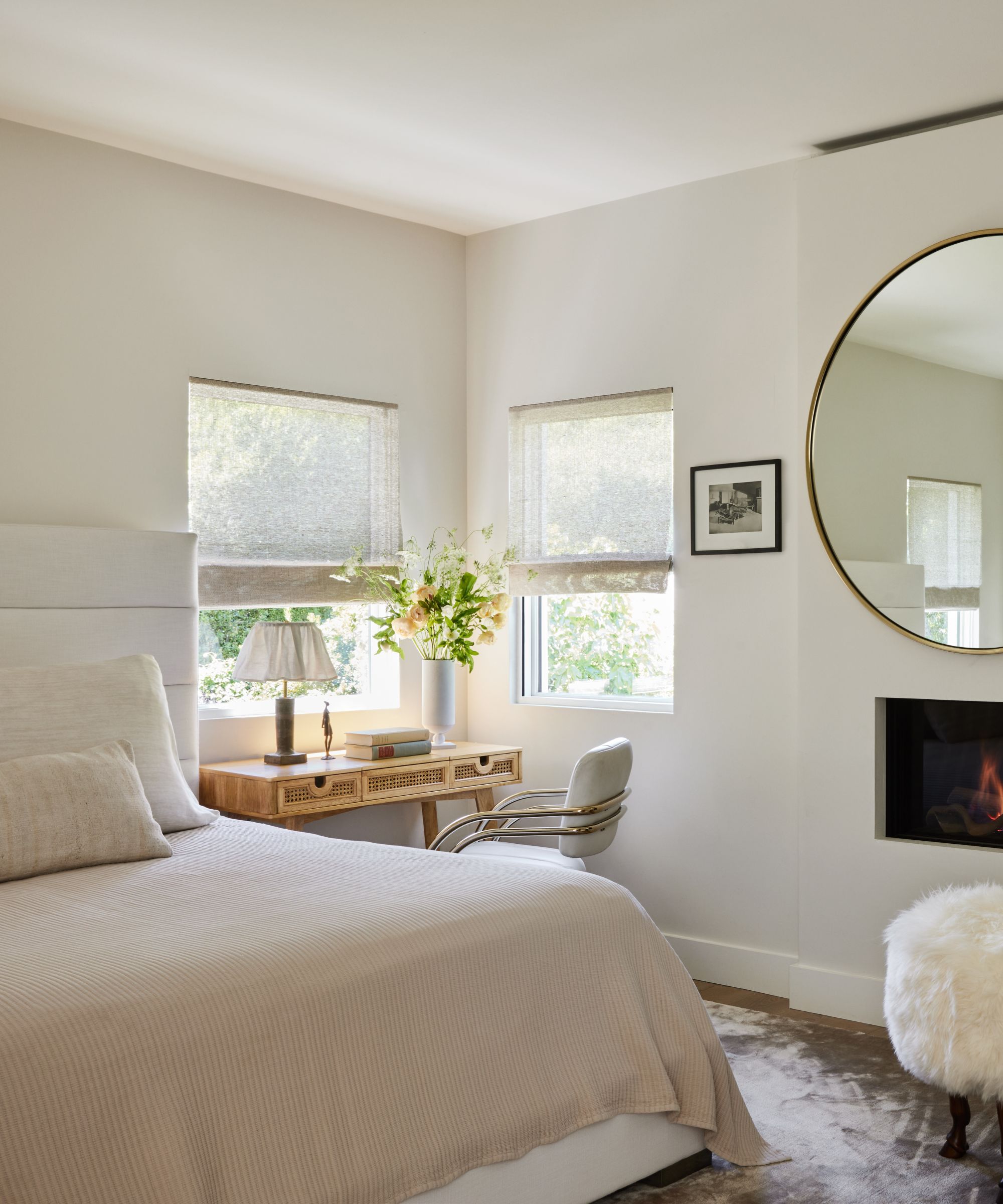 A calming, neutral bedroom with warm white walls, neutral bedding, and a desk in the corner next to two windows.
