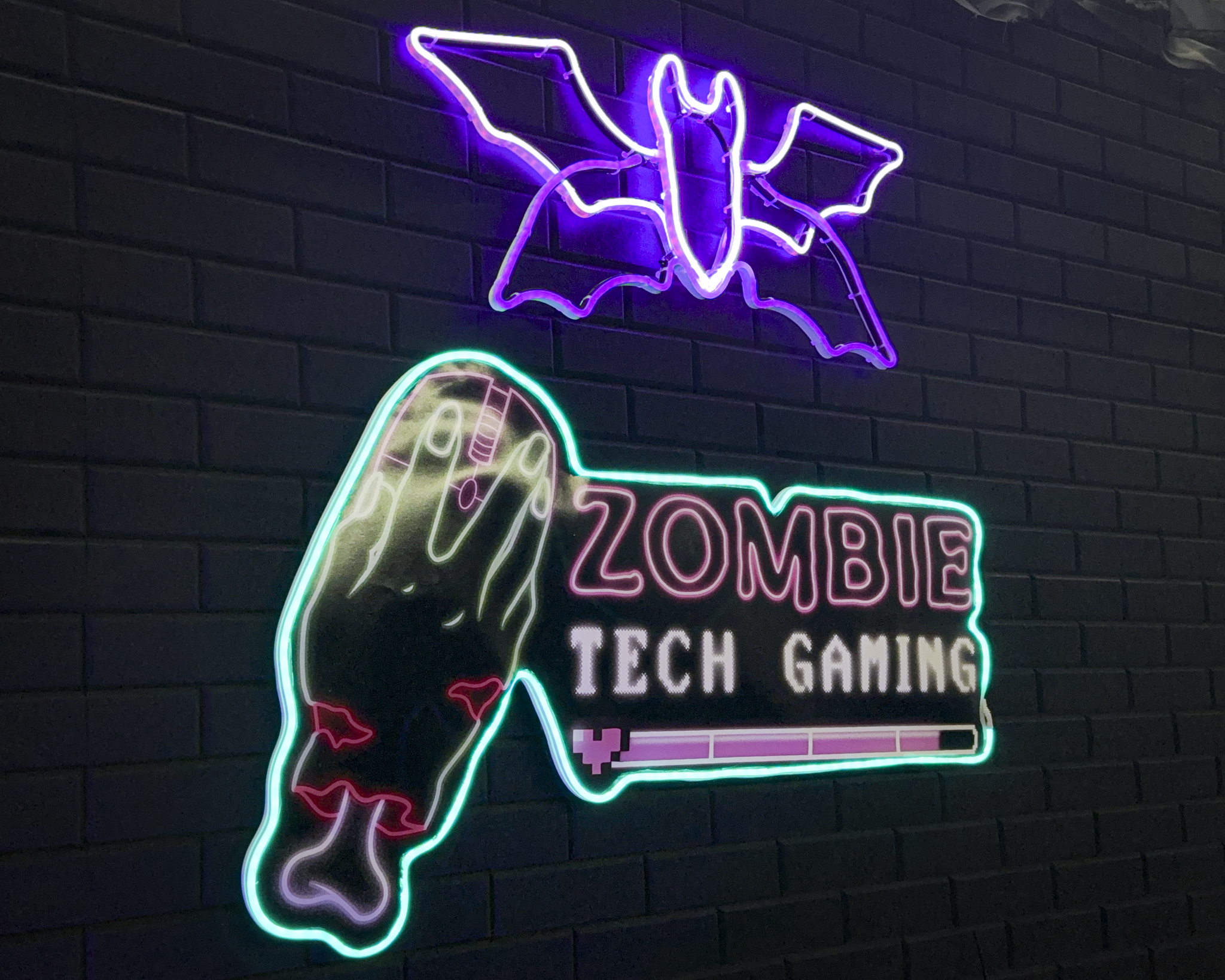 The Zombie Tech Gaming logo outlined in neon, and a bath neon light asbove it. Both are mounted on a brick wall painted black.