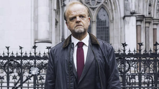 British actor Toby Jones, dressed in a sombre grey suit, stands outside the High Court in London, in a scene from ITV drama Mr Bates vs The Post Office