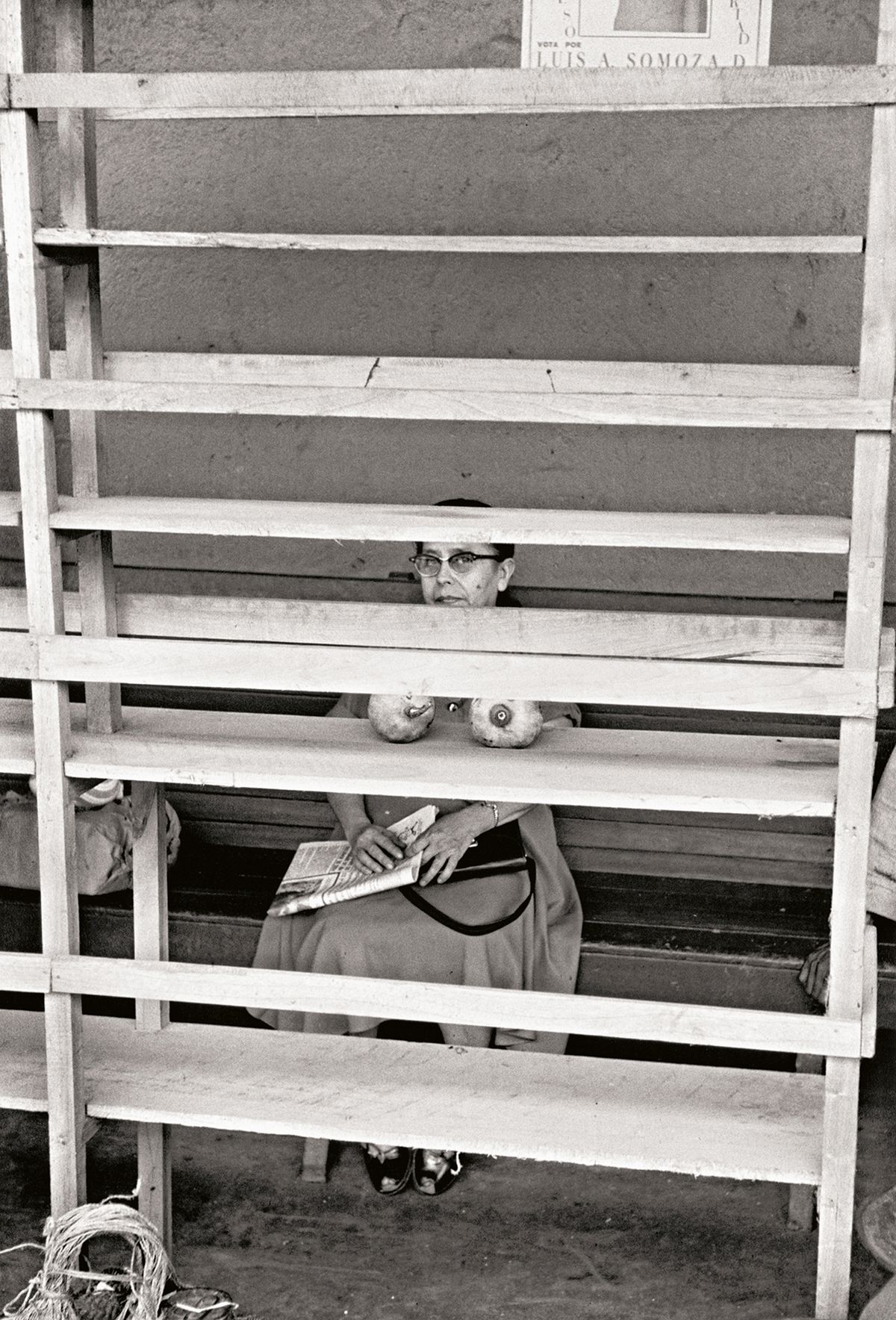 Black and white image of a woman in a dress sits behind wooden slats, holding a newspaper in her lap, with a bag beside her