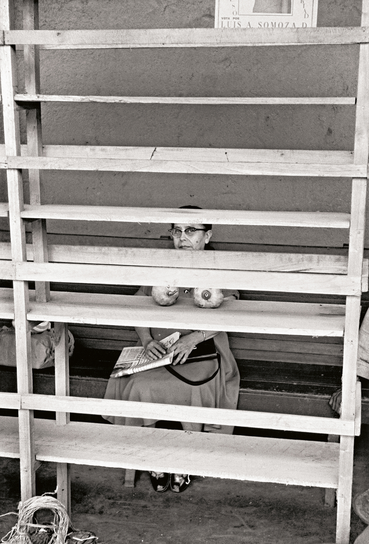 Black and white image of a woman in a dress sits behind wooden slats, holding a newspaper in her lap, with a bag beside her