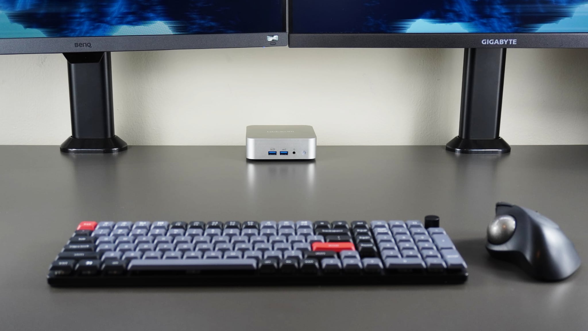 The Geekom A6 at the back of a desk under two monitors with a mechanical keyboard and trackball mouse in the foreground