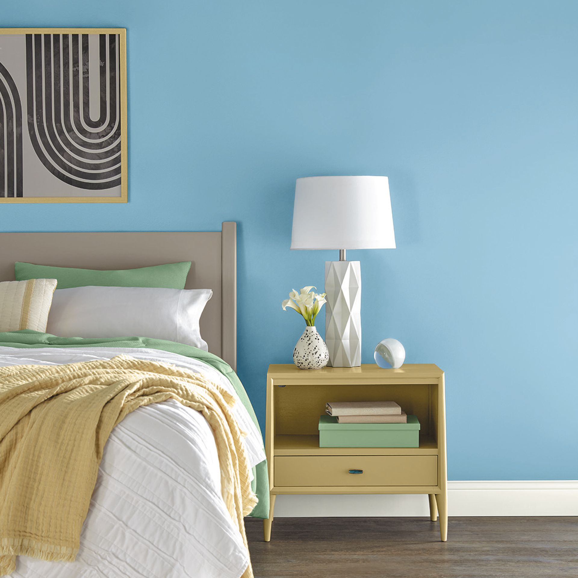 Dinding biru langit di interior kamar tidur modern dengan meja dan lampu samping tempat tidur kuning, seprai musim semi yang segar, dan cetakan seni berbingkai di atas kepala tempat tidur.