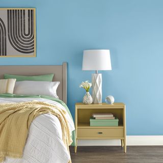 Sky blue wall in modern bedroom scheme, with yellow bedside table and lamp, spring fresh bedlinen, and curves framed art print above headboard.