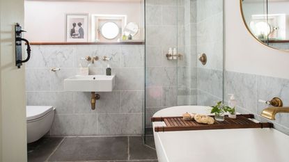 Bathroom with white bath, glass shower cubicle, basin and toilet, grey wall tiles and pale grey walls.