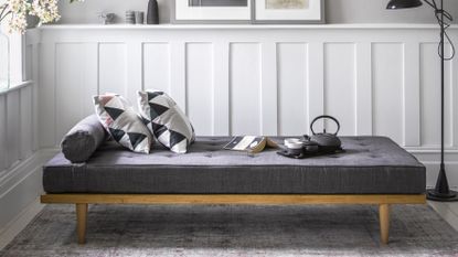 a large grey day bed – one of the best guest beds – with cushions, a book and a kettle on top of it, in a grey living room, with white panelling on the walls, artwork propped up against the wall, a black floor lamp, and a window to the left