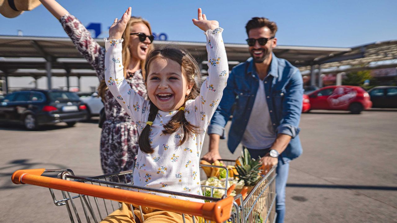 A little girl sits in a shopping cart pushed by her parents.