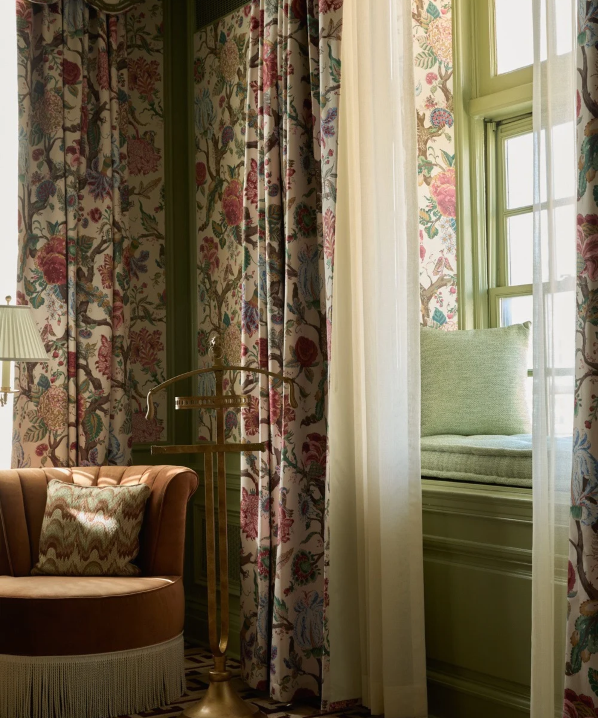 Chintz-drenched bedroom corner, featuring floral-patterned wallpaper, drapes, and a fringe accent chair