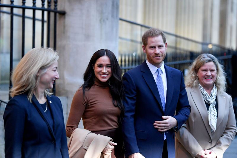 Meghan Markle wears a brown sweater and holds hands with Prince Harry who wears a navy suit with a white shirt