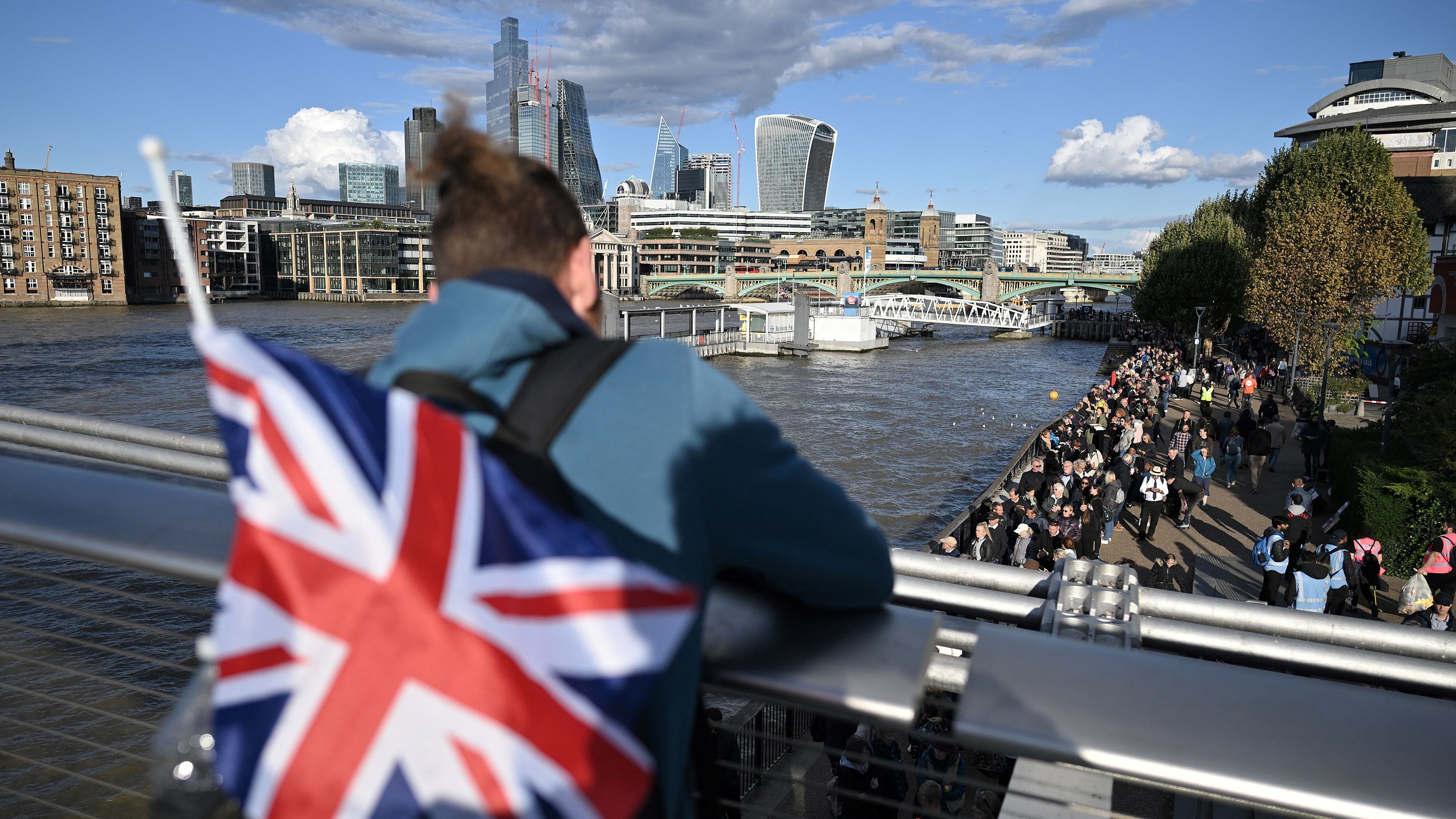 man with british flag standing on bridge looking at lineup of people