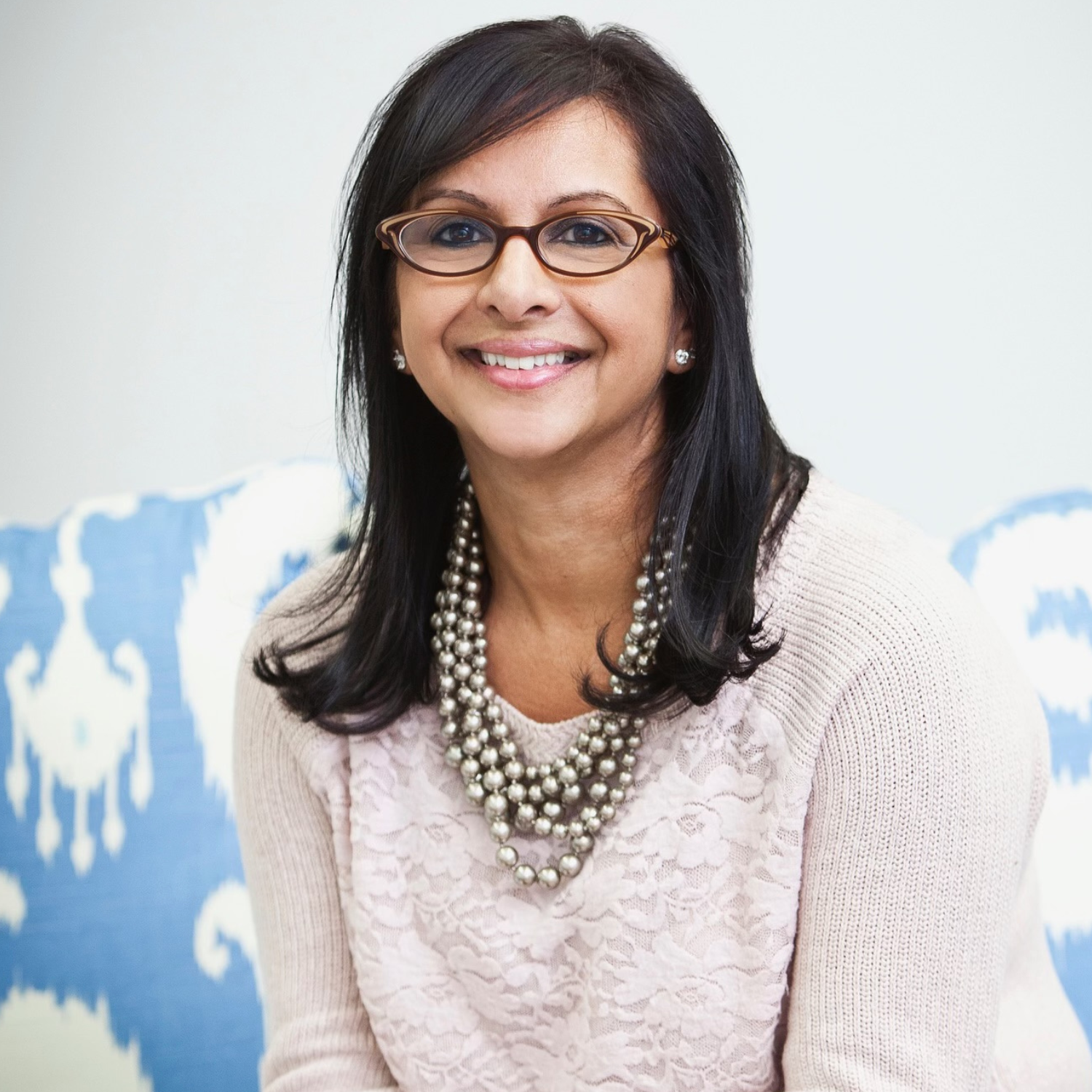 A woman with short black hair in brown rimmed glasses and a lace and knit sweater with strands of pearls