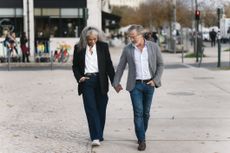A stylish senior couple is holding hands and walking down a sidewalk in a European city.