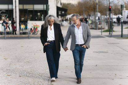 A stylish senior couple is holding hands and walking down a sidewalk in a European city.