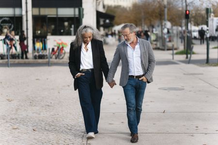 A stylish senior couple is holding hands and walking down a sidewalk in a European city.