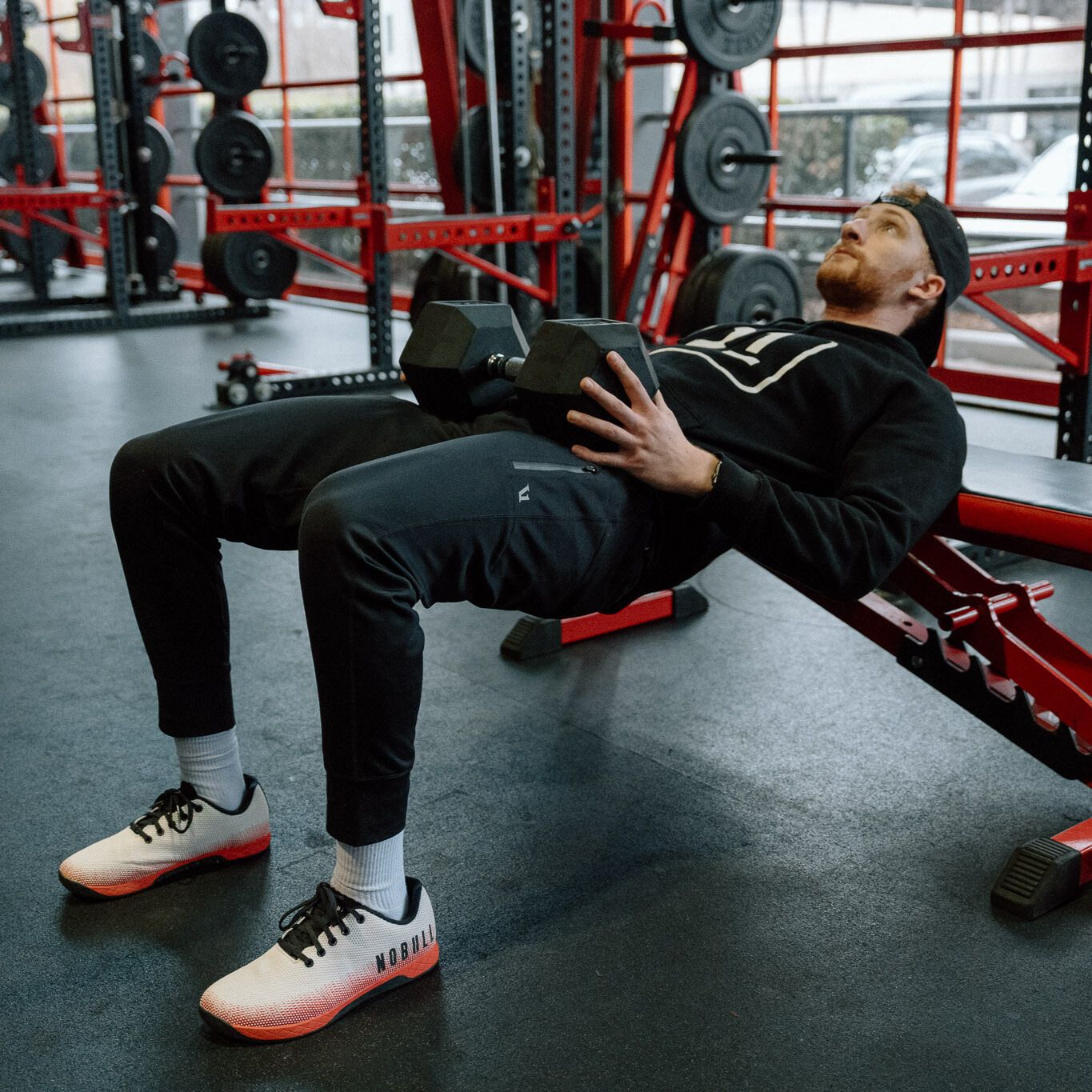 man dressed in black trousers and hoodie performing a hip thrust on a gym bench with a dumbbell on his hips, sideways to the camera