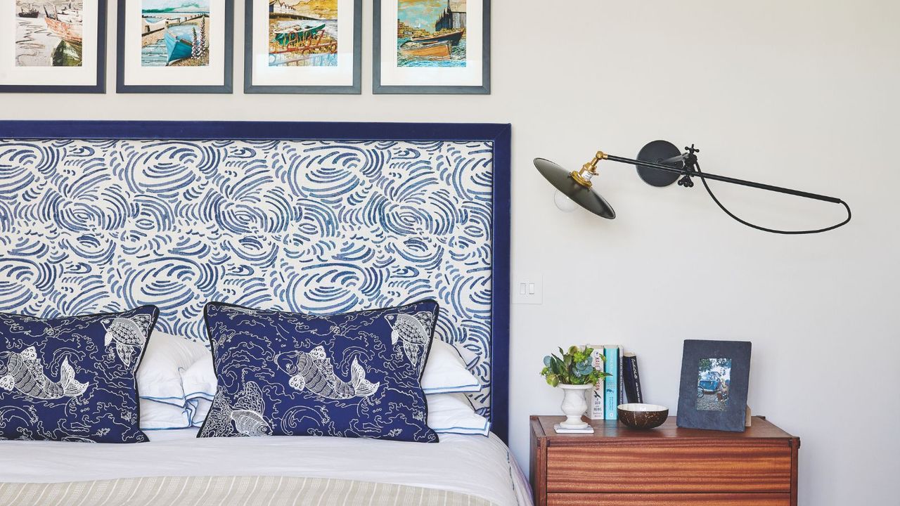 A white bedroom with a blue water patterned headboard bed with blue koi fish cushions and bedding, framed colorful images above, a wooden beside table with books, a plant, and a framed photograph in a matching blue frame and an industrial style moveable lamp above