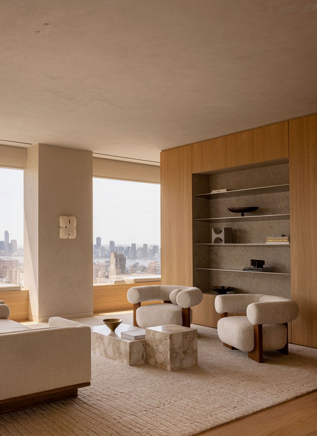 Living room at 262 Fifth Avenue with sculptural upholstered armchairs, marble tables, oak cabinetry, and built-in stone shelving overlooking Manhattan