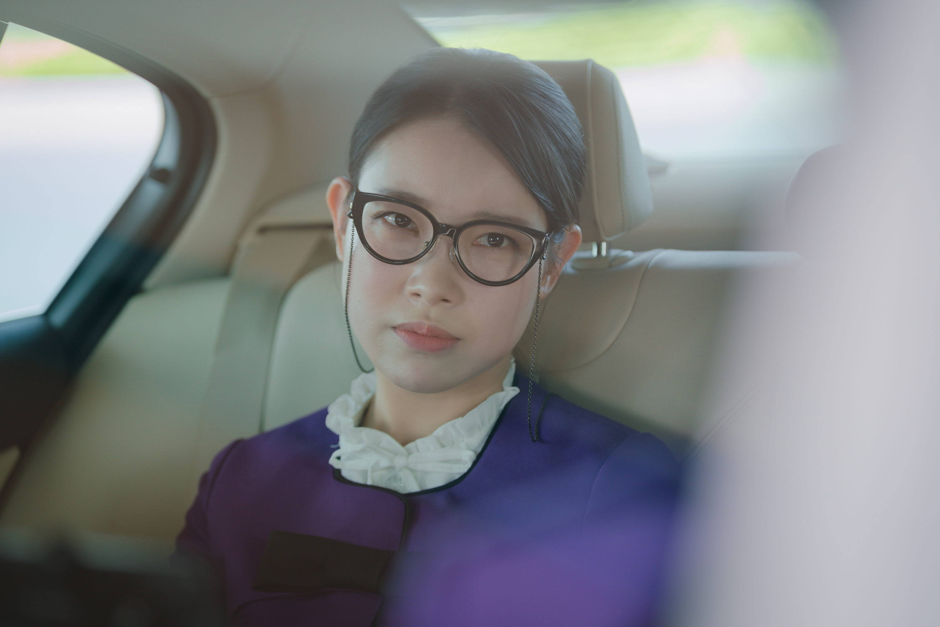 A woman (Kim Ah-young as Choi Min-ju) wears a purple cardigan, a white frilly shirt, and glasses as she sits in the backseat of a car, in a still from the Netflix K-drama 'Boyfriend on Demand.'