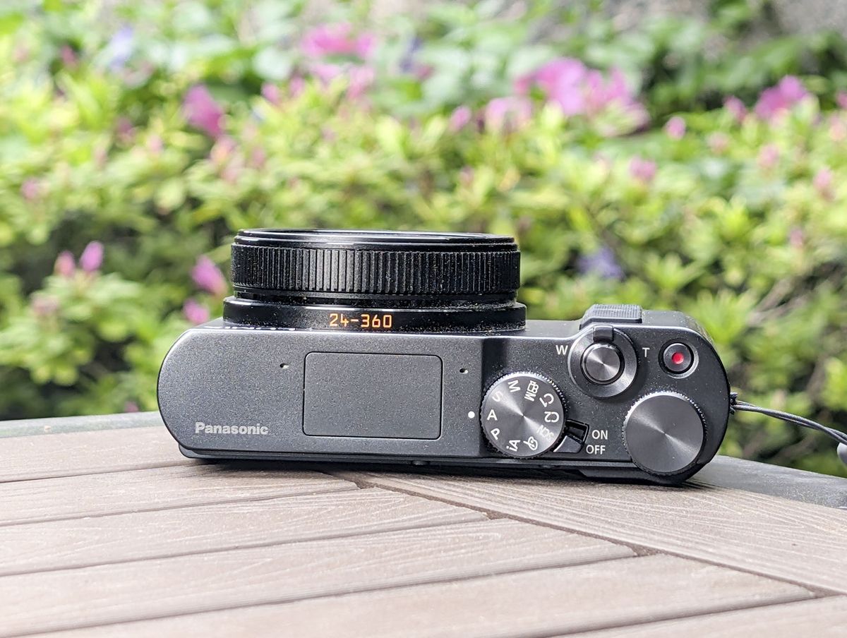 The top plate of the Panasonic Lumix TZ300 compact on a wooden table, with lens packed away, and a bibrant floral backdrop