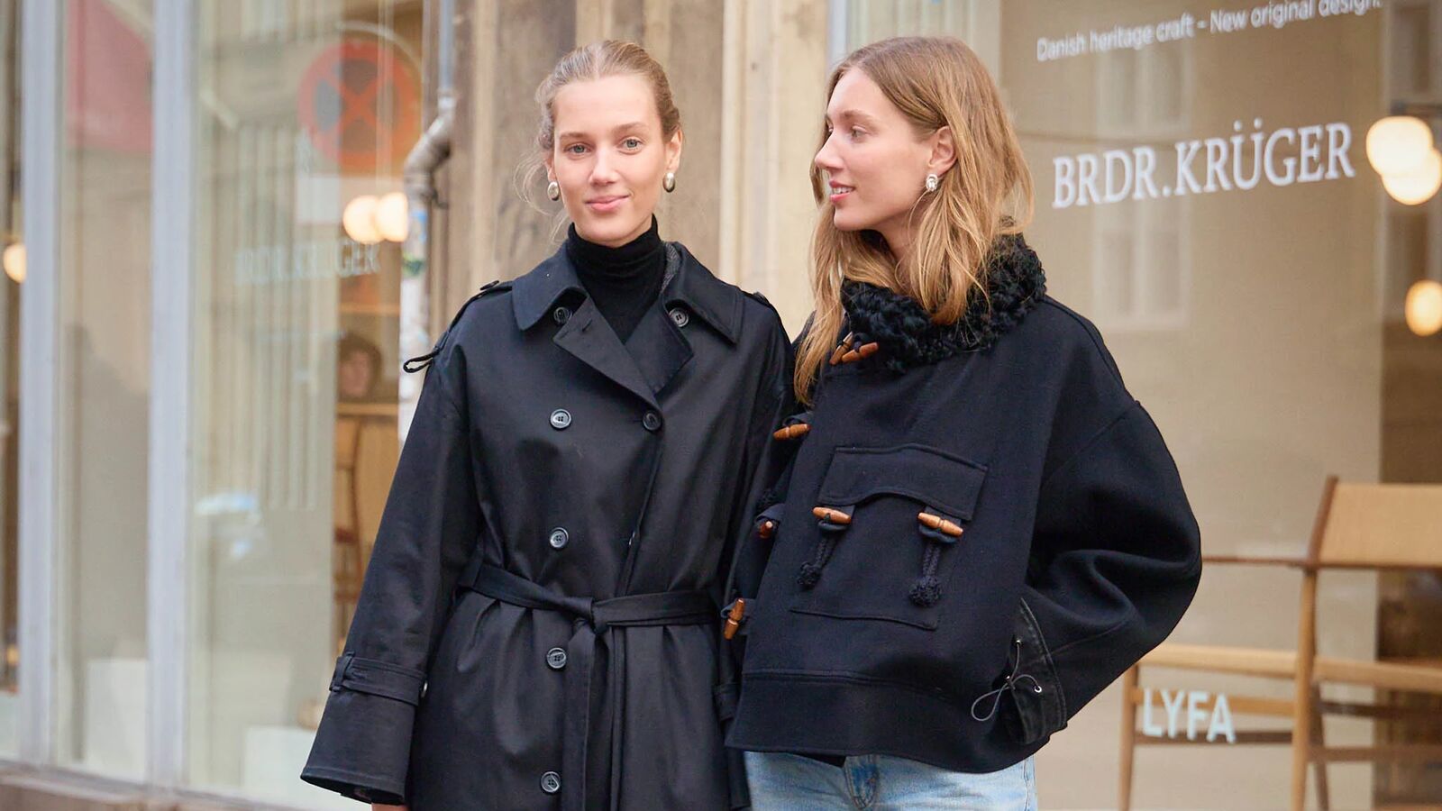 Two women are posing for street style photos in Copenhagen. One woman is wearing a is wearing a black duster trench style coat. The woman is wearing a short cropped coat and jeans. 