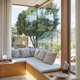 a glazed corner window seat with cushions overlooking a garden with a greenhouse