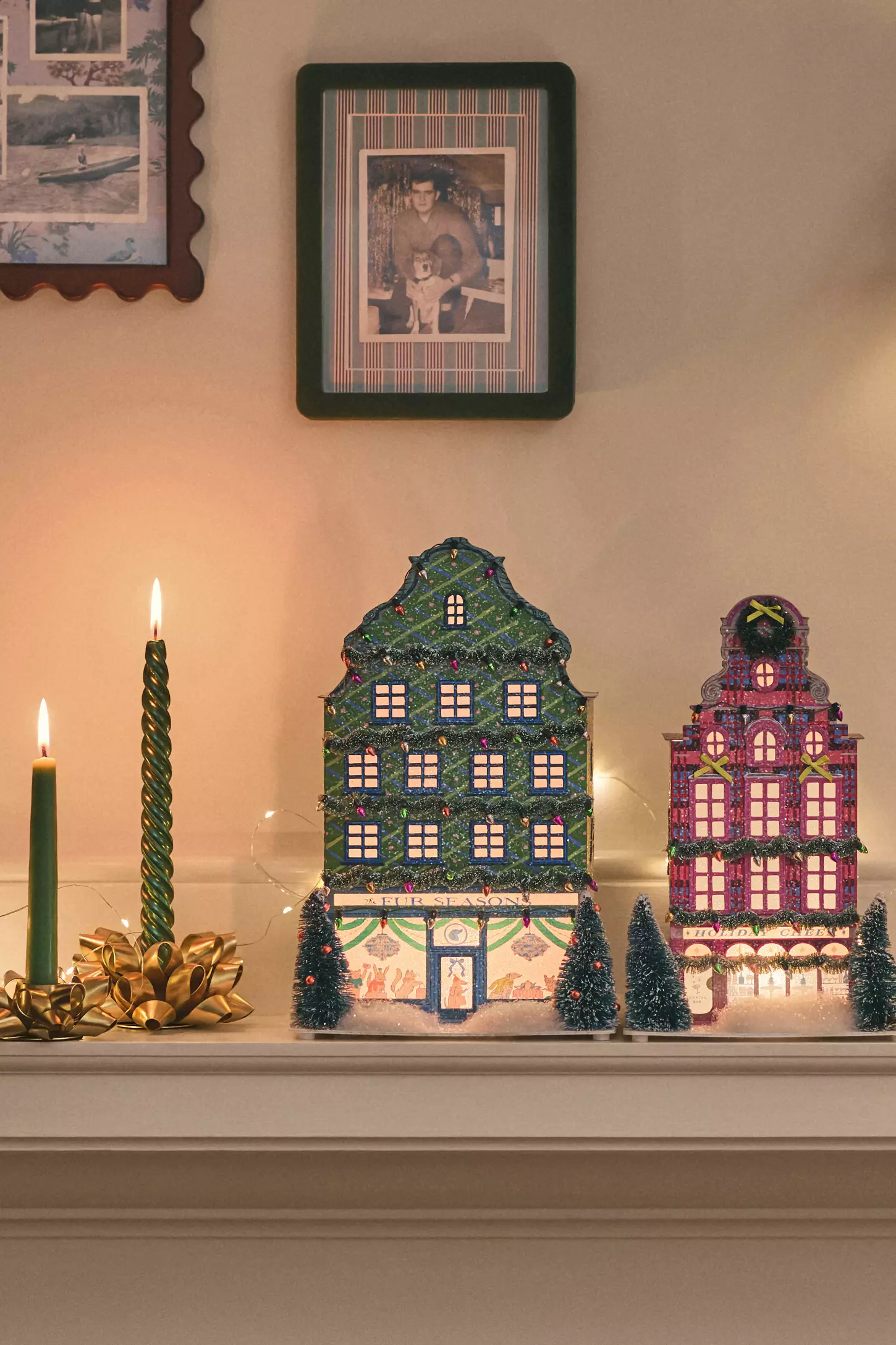 Two glittery and colorful paper light-up christmas houses on a mantel next to two lit green taper candles with framed pictures on the wall above them