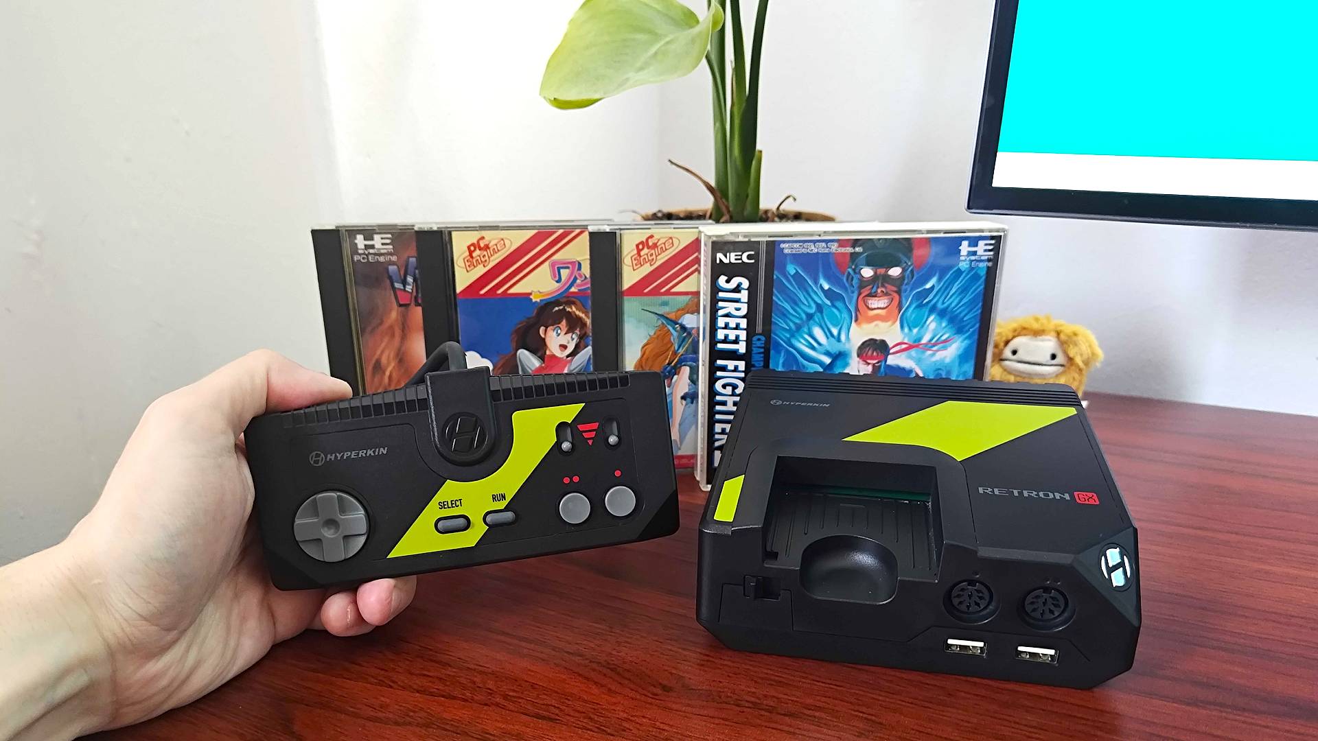 Hand holding Hyperkin Retron GX controller next to console with PC Engine games in backdrop next to plant.