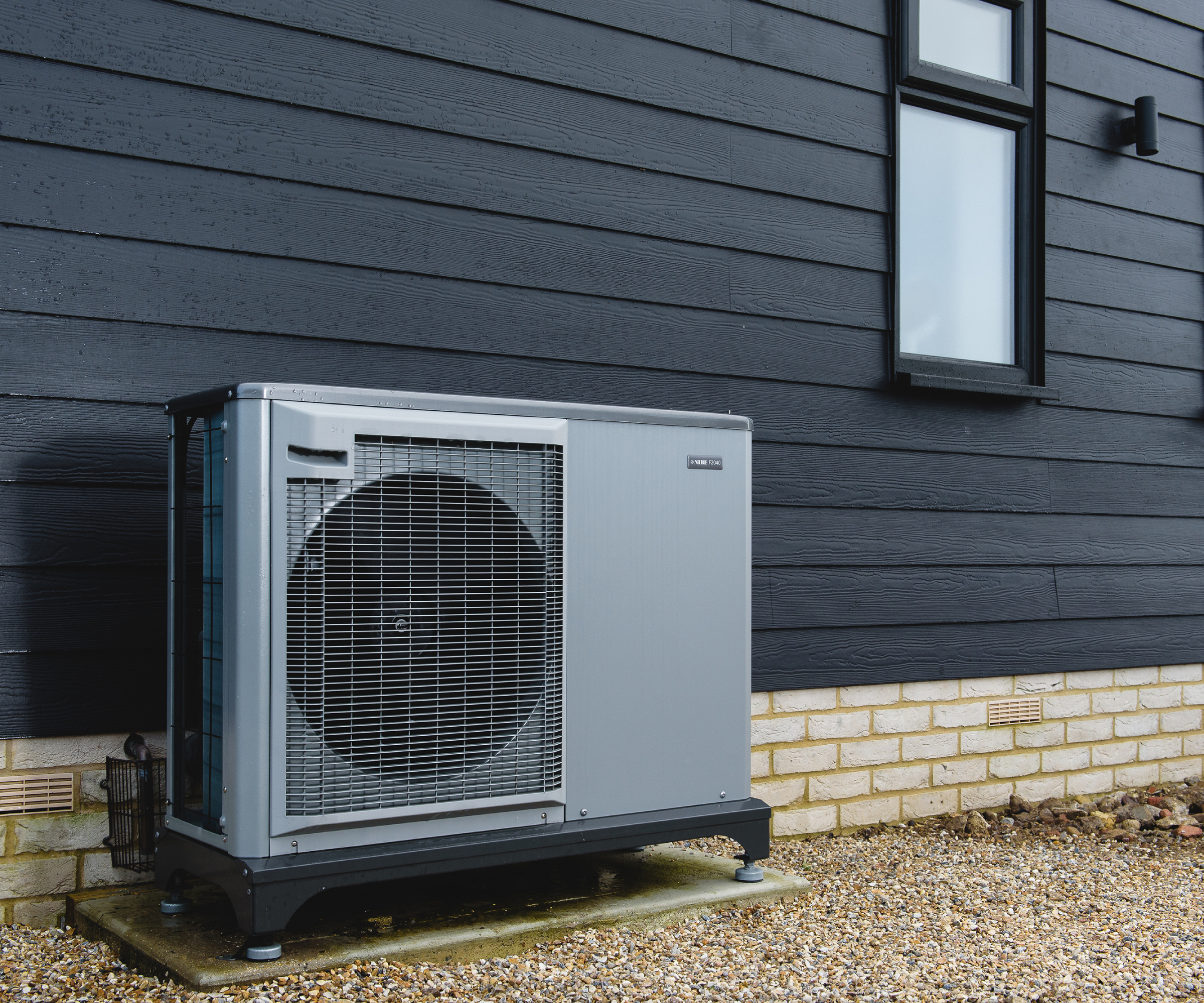 grey air source heat pump against black clad house with lower section in pale brick and gravel path