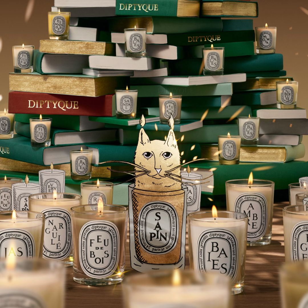 Diptyque holiday collection 2025 imagery with stacked books, lit candles, and illustrated cat image