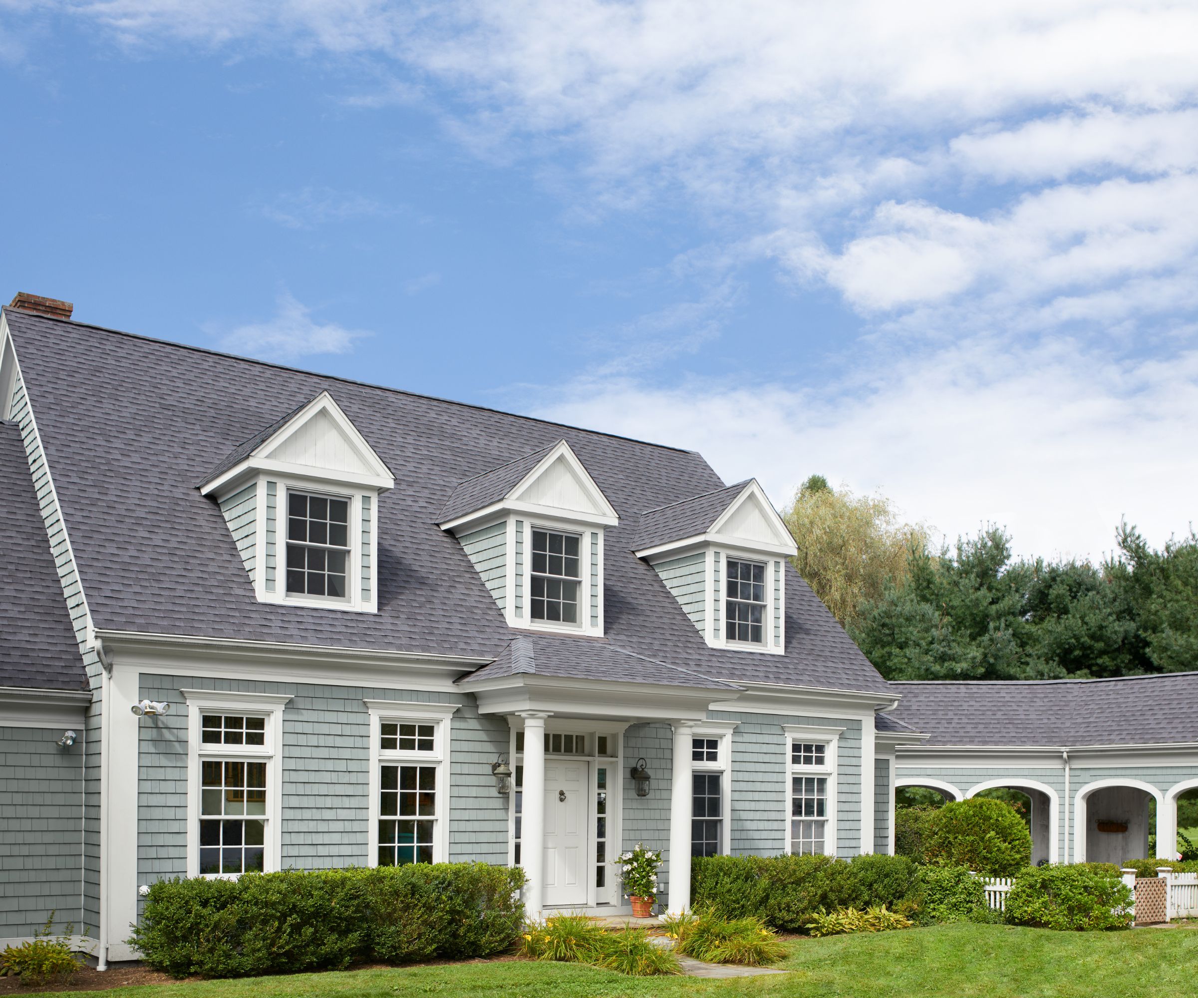 Colonial-style house with Boothbay Gray painted exterior, white front porch with pillars and white painted front door, and grassy front lawn with hedges surrounding the house.