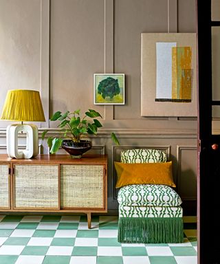 Entryway with green and white checked floor, green pattered lounge chair with fringing detail, dark wood and wicker cabinet, light brown painted paneled walls, artwork, decorative ornaments and table lamp on sideboard