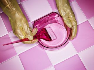 A pair of hands, wearing gloves are mixing hair dye in a pink bowl with a pink brush. The background is checkered pink.