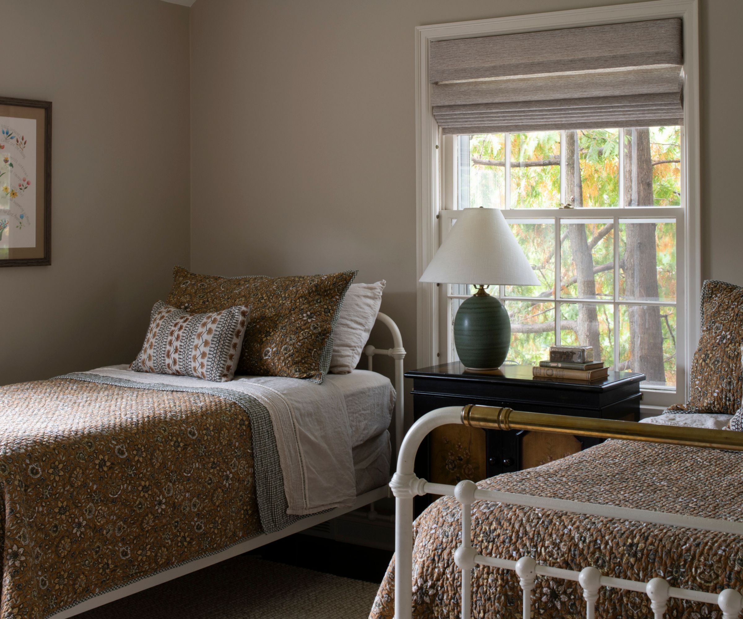 twin bedroom with neutral walls, two white wrought iron single beds styled with vintage yellow floral bedspreads with patchwork throw pillows and a chest with a green table lamp