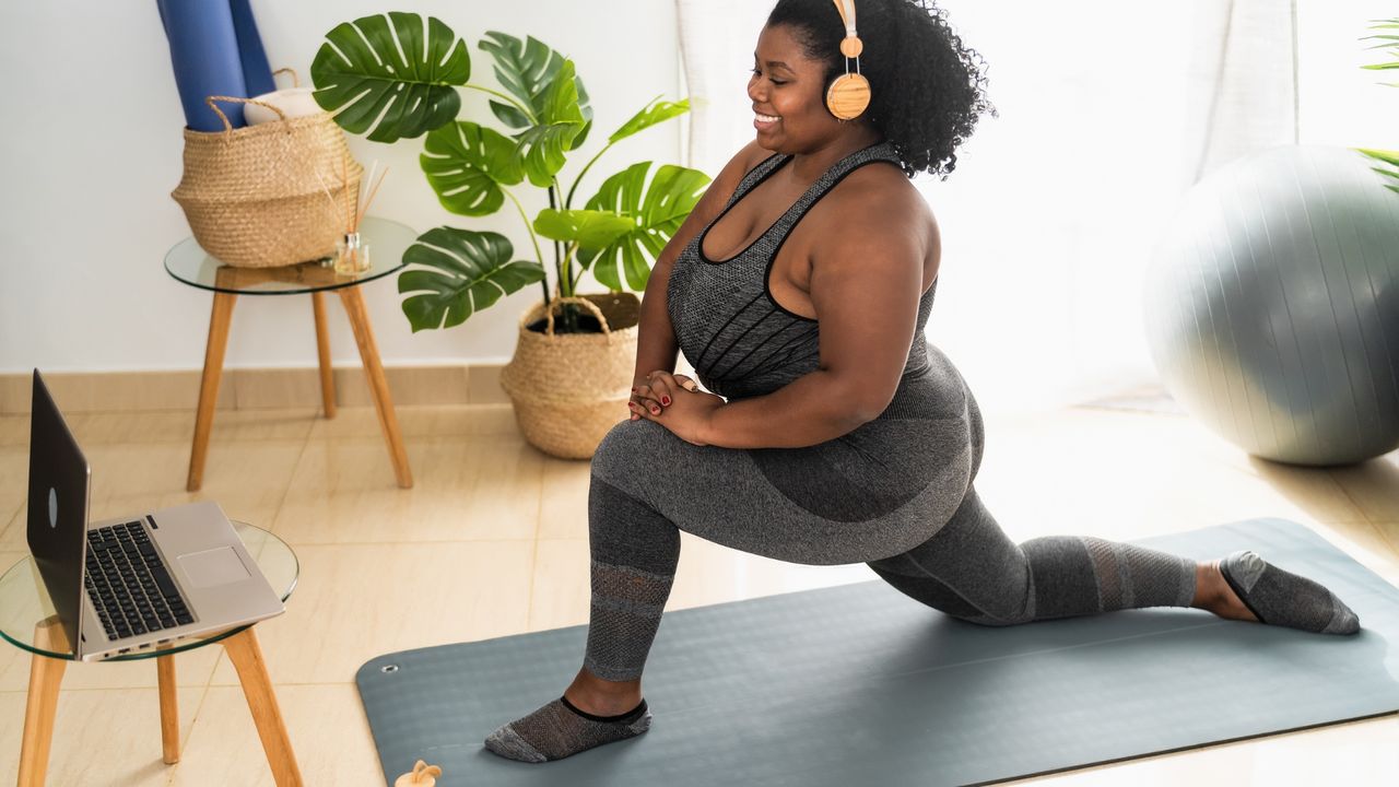 black woman afro hair yellow headphones grey leggings crop top low lunge facing laptop plants balance ball background