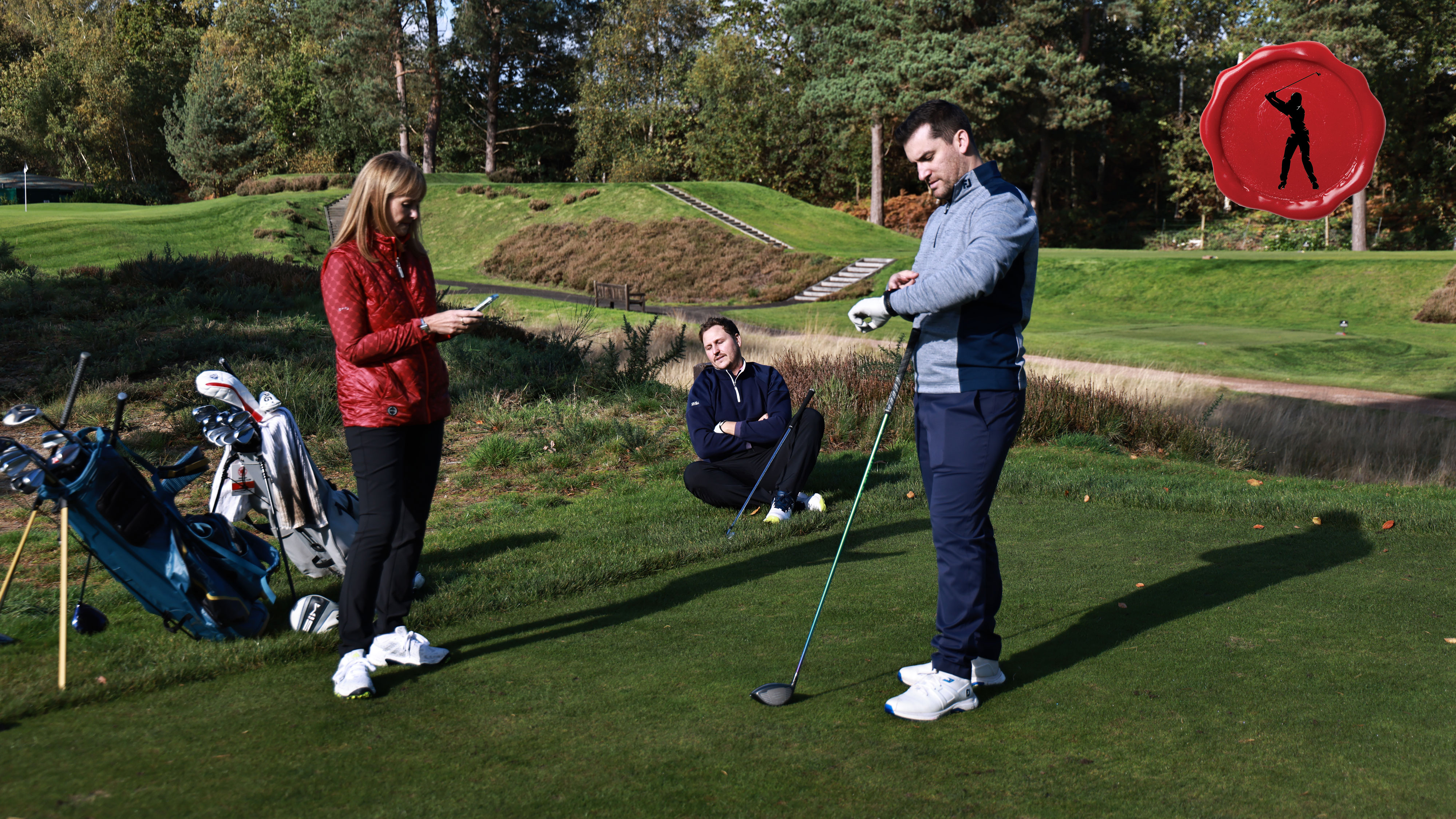 three golfers waiting on the tee because of slow play, with one sitting on the ground, one looking at their phone and one looking at their watch