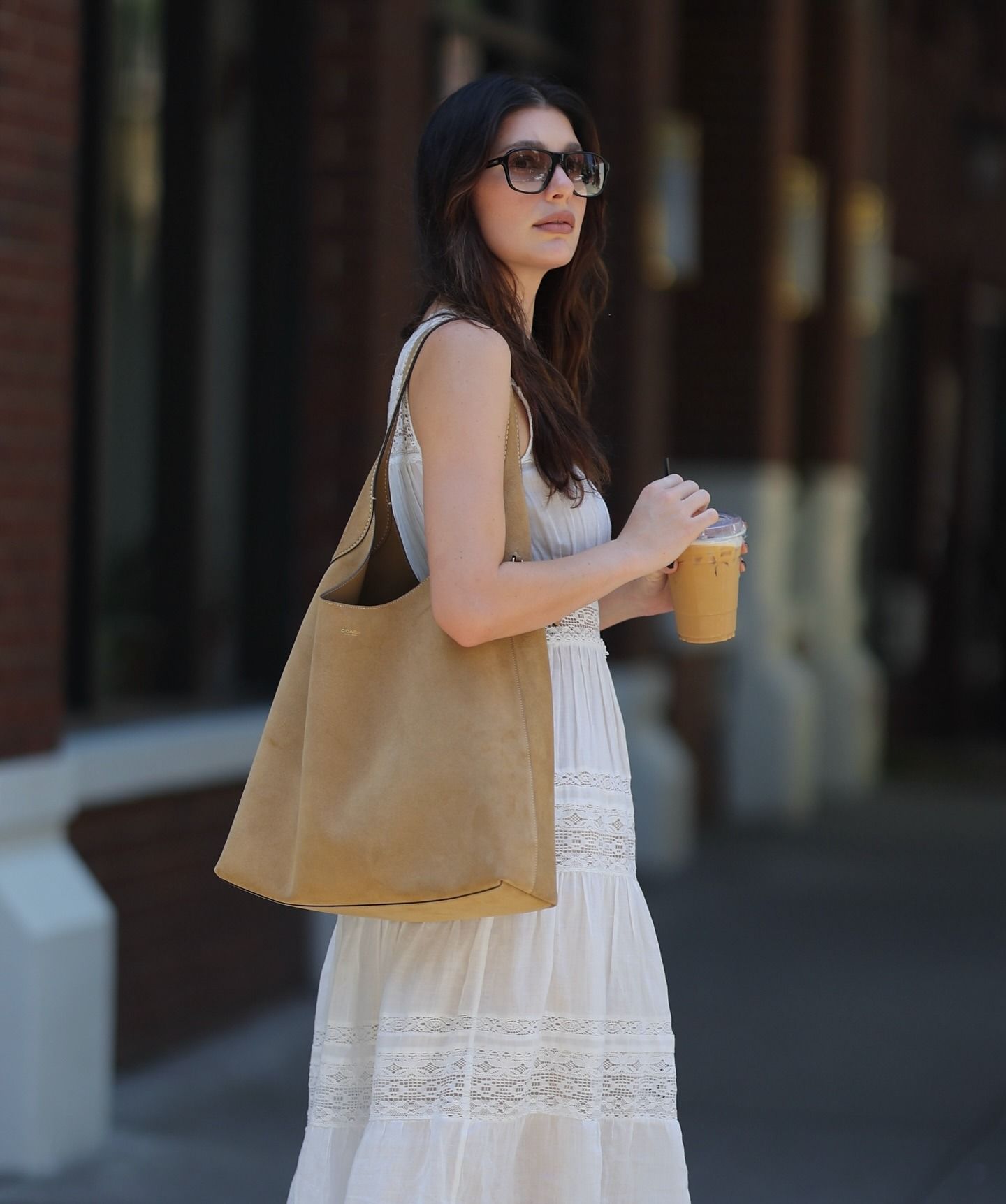 Camila Morrone is wearing a a white tiered lace prairie dress, light brown cowboy boots and carrying a beige coach Brooklyn Shoulder bag in 28. She in pictured walking down the streets of NYC in a recent image.