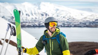 A skier holds his skis and chats to a friend with a lake in the background