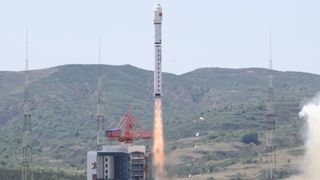 a white chinese long march 2d rocket launches into a pale blue sky with greenish-brown mountains in the background