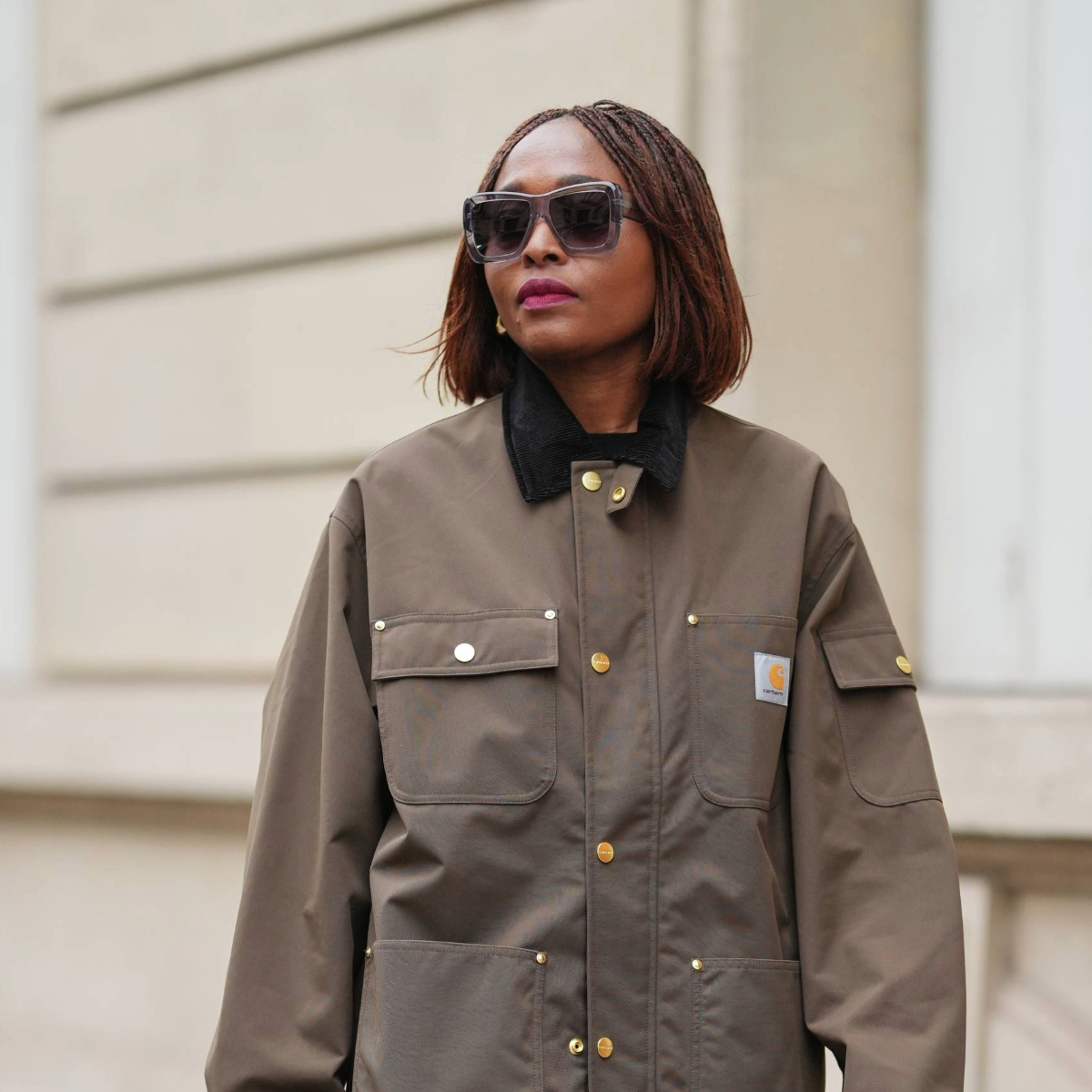 PARIS, FRANCE - DECEMBER 13: Carrole Sagba wears a straight chin-length bob hairstyle with a center part, makeup featuring a natural complexion with soft pink lipstick, an olive green khaki cotton canvas workwear jacket with flap chest pockets and snap-button fastening by Carhartt, black opaque tights, a burgundy leather tote bag with a structured silhouette and crocodile pattern, and burgundy shiny knee-high leather boots with a pointed toe and block heel and crocodile pattern, during a street style fashion photo session, on December 13, 2025 in Paris, France. (Photo by Edward Berthelot/Getty Images)