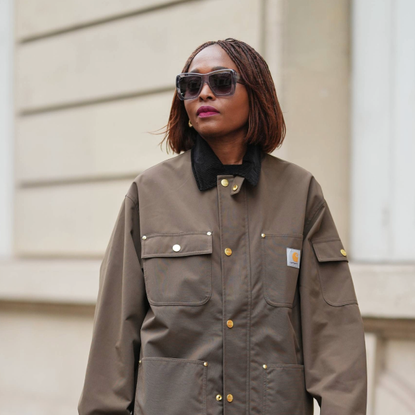 PARIS, FRANCE - DECEMBER 13: Carrole Sagba wears a straight chin-length bob hairstyle with a center part, makeup featuring a natural complexion with soft pink lipstick, an olive green khaki cotton canvas workwear jacket with flap chest pockets and snap-button fastening by Carhartt, black opaque tights, a burgundy leather tote bag with a structured silhouette and crocodile pattern, and burgundy shiny knee-high leather boots with a pointed toe and block heel and crocodile pattern, during a street style fashion photo session, on December 13, 2025 in Paris, France. (Photo by Edward Berthelot/Getty Images)