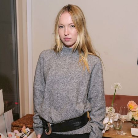 Lila Moss poses for a photo wearing black capri pants with a grey jumper, an oversized belt and black slingback heels.