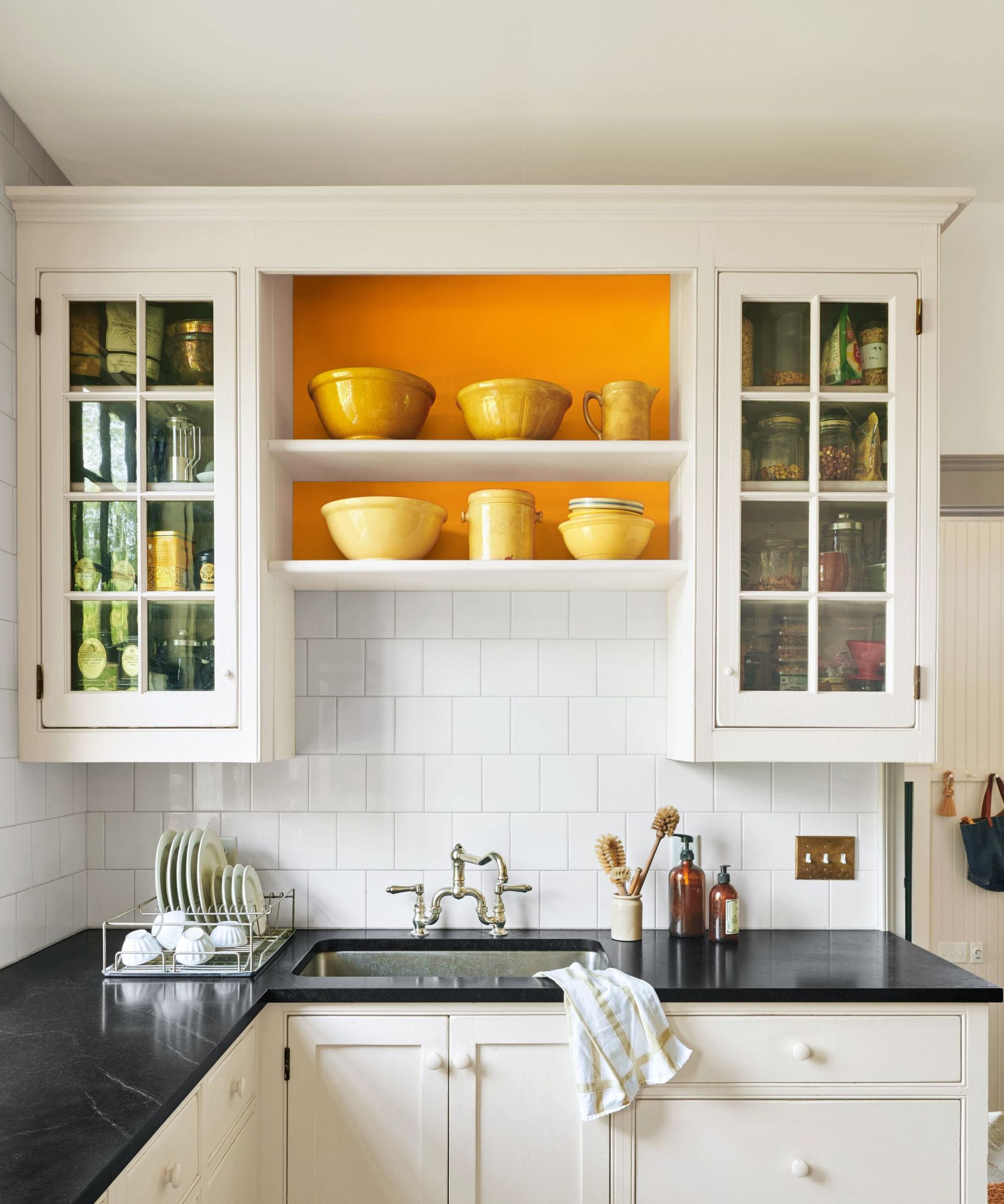 A kitchen with white cabinets, white wall tiles, black countertops, and the back of open shelving painted bright orange