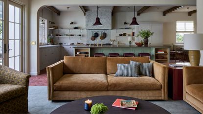 a large open plan living and kitchen space, taken from the seating area showing a tan cord sofa and round coffee table styled on terracotta floors looking through to a layered gray and green kitchen
