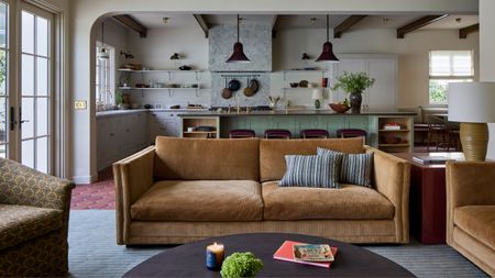 a large open plan living and kitchen space, taken from the seating area showing a tan cord sofa and round coffee table styled on terracotta floors looking through to a layered gray and green kitchen