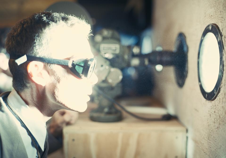a man (cillian murphy as j robert oppenheimer) wearing goggles looks through a circular window as a bright light flashes across his face, in 'oppenheimer'