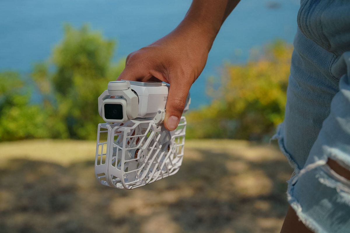 HoverAir Versa 2-in-1 drone and pocket gimbal camera in white, with the wings folded down, in person's hand by a dramatic coastline on a sunny day