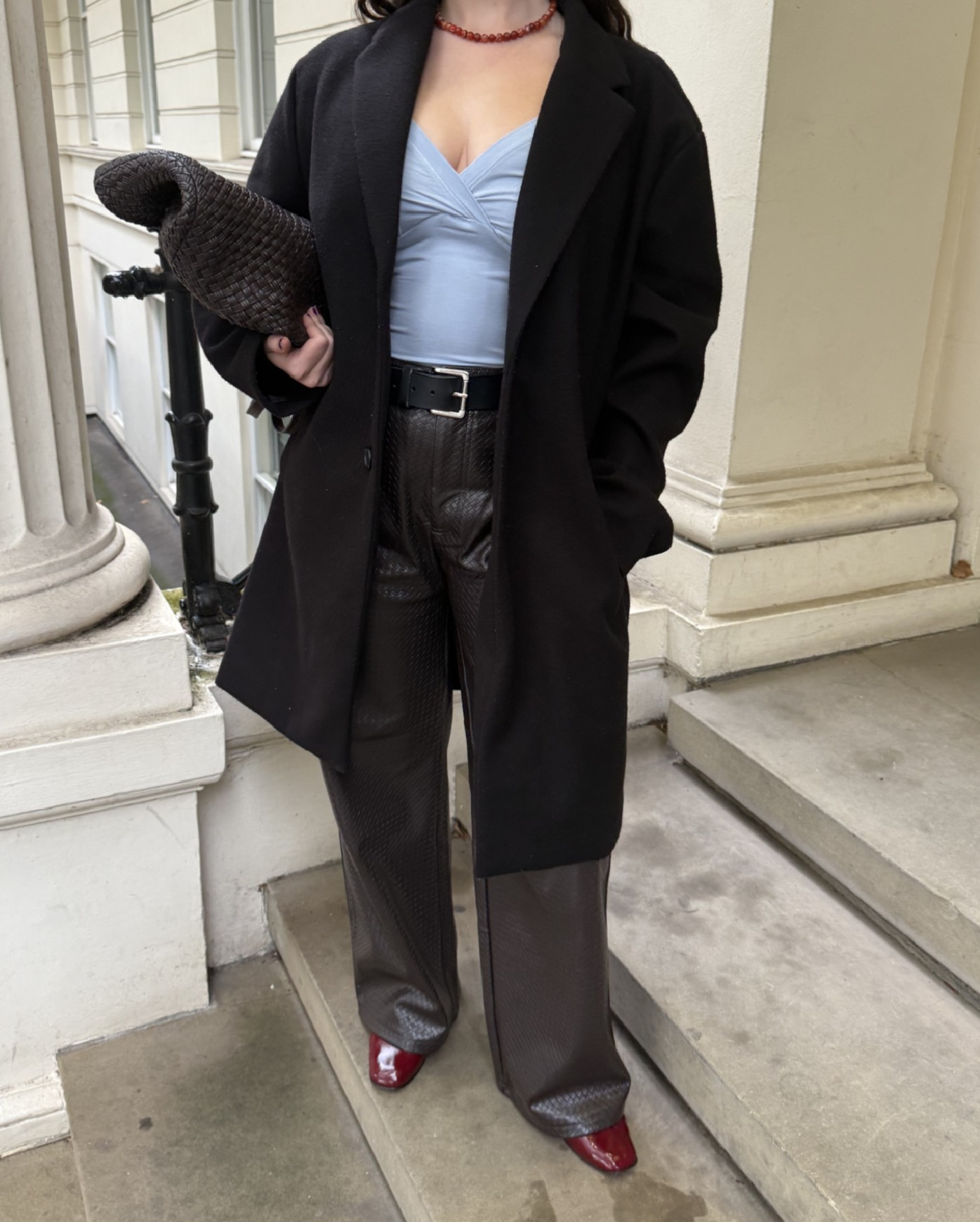 An image of Who What Wear&#039;s Ava Gilchrist wearing woven leather trousers with a woven leather bag, red ballet flats, a blue top from Mode Mischief and a black coat.